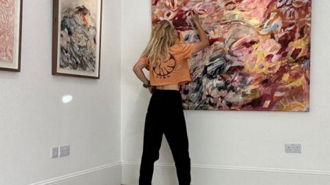 Artist Emma Howell working on a painting that is mounted on the wall. She has long blonde curly hair and wears an orange cropped top and black trousers.