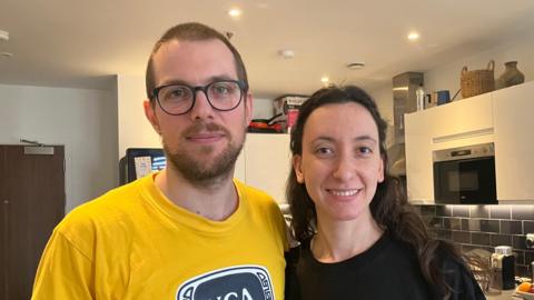 A man in a yellow top stands next to a woman in a black one. He has short brown hair and glasses. She has long dark brown hair