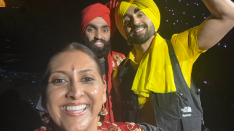 The couple are standing with Diljit on stage and all are smiling at the camera in this close-up selfie. Diljit has a yellow and black outfit on and yellow turban. Priptal also has a red turban on.