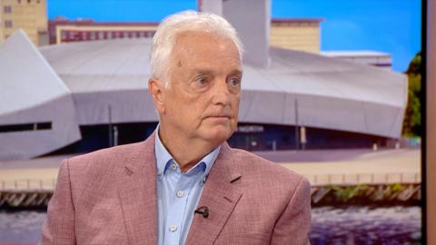A man in a pink suit speaks in a studio.