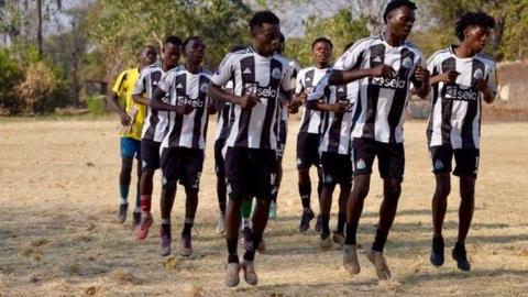 A group of young African men jumping up and down as part of a training exercise. They are all wearing black and white Newcastle United kits. The ground is grassy and dusty and there are trees in the background.