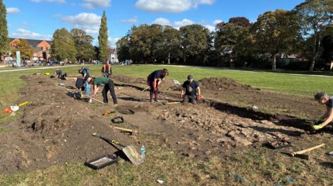 A wide view of the park where a huge pit about a foot deep has been dug. The soil removed from it lies on either side. Various people are bent down looking into the pit.