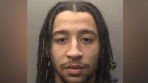 Head-and-shoulders custody photograph of Tristan Oliver with long braided hair, facing the camera against a plain grey background. He is wearing a dark zip-up top.