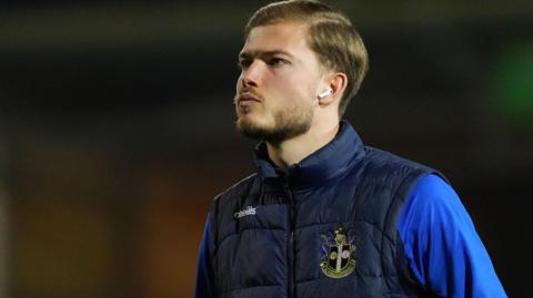 Lewis Simper wearing a blue Sutton top looks to his right with a straight face