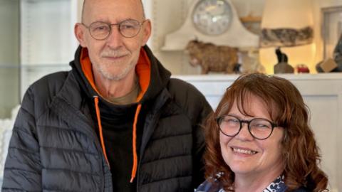 Rob Sim, who has a grey beard and is wearing glasses, and Dawn Sim, who has long wavy brown hair and glasses.