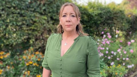 A woman in green stands against a flower and hedge backdrop