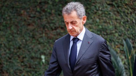 Sarkozy, wearing a navy suit, looks down as he walks beside a big green bush.