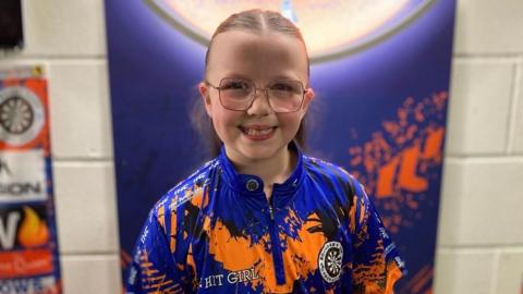 Heidi smiling at the camera, with her long brown hair tied back, wearing pink metal-rimmed glasses and a blue and orange Boundary Park Darts top.