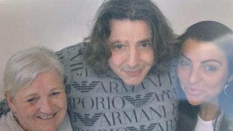 A middle-aged man with long dark hair stands with has arms around a younger and an older woman.