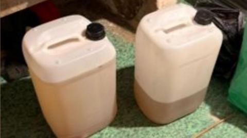 Two plastic containers filled with brown liquid. The containers are on a green carpet surrounded by cardboard boxes.