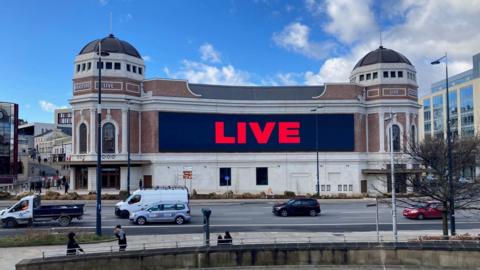 A red-brick building with two turrets at either end and a giant display screen showing the word 'live' in red letters.