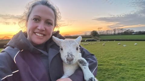 A person is taking a selfie while holding a young lamb in a grassy field at sunset. The lamb is white with dark markings on its nose and legs, and in the background there are several sheep scattered across green pasture, hedgerows, trees and a colourful orange and blue evening sky stretching across the horizon.