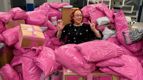 Rachel Grady sits among a large pile of pink parcels. She has a ginger curled bob and holds two peace signs up to either side of her head. She smiles widely and wears pink glasses.