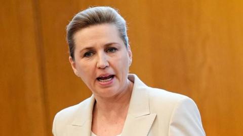 Mette Frederiksen speaking in parliament, wearing a beige jacket over a white top