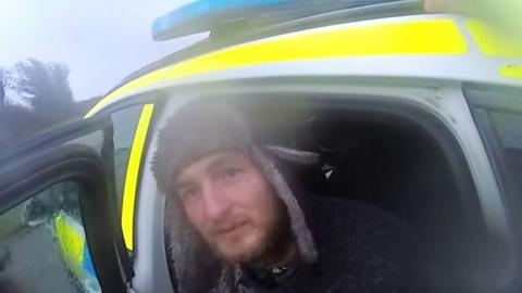 A grab from police body worn camera footage showing Alexander Lewis-Ranwell sitting with his head out of the back door of a police car. The yellow and blue livery of the police vehicle is partially visible. Lewis-Ranwell is looking towards the camera and is wearing a dark coloured hat with light coloured fleece covering the forehead area and ear flaps.