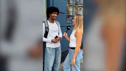 A man and a woman talking, both wearing blue jeans and white tops, in a street. She holds a microphone.