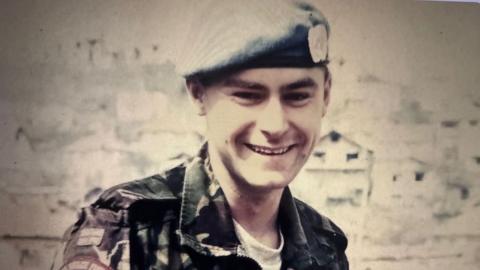 A picture of a young soldier smiling; he is wearing a beret and military greens. In the background, faint outlines of houses can be seen.
