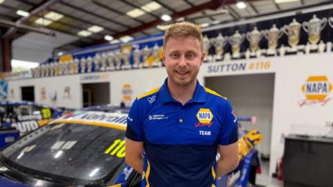 Ash Sutton in a blue polo shirt standing in a garage with a racing car behind him.