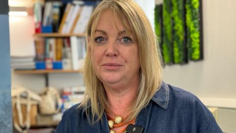 a woman with shoulder length blond hair is looking into the camera. She's sitting in an office and is dressed in a denim shirt.