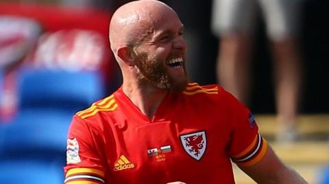 Footballer Jonny Williams looking to his left and laughing to celebrate a goal as a player for the Wales national football team while wearing a red shirt with yellow trim and the Wales and Adidas logos on the chest in a stadium