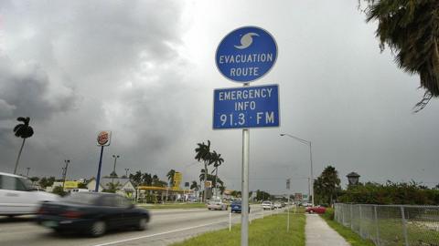 Photo shows a road with cars passing a blue sign that has a hurricane symbol and says 'evacuation route' 'emergency info 91.3FM'