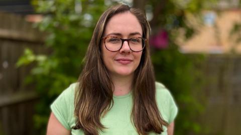 Erin Cooper wearing a green top and glasses. She is looking directly at the camera and standing in a garden. She has long brown hair hanging to chest level.