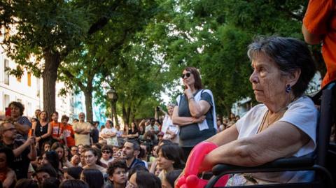 Maricarmen sits outdoors under some trees in a wheelchair. Behind her are a crowd of protesters