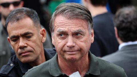 Former Brazilian President Jair Bolsonaro wearing green polo shirt with police officer standing behind him