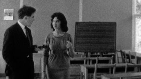 Black and white image of a teacher in an empty classroom talking to a BBC reporter.