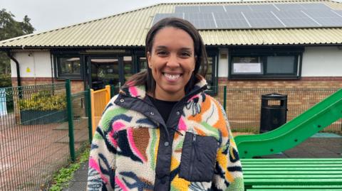 Fahren Peach, a woman with dark hair wearing a colourful, patterned fleece, is stood in a playground with a building in the background.