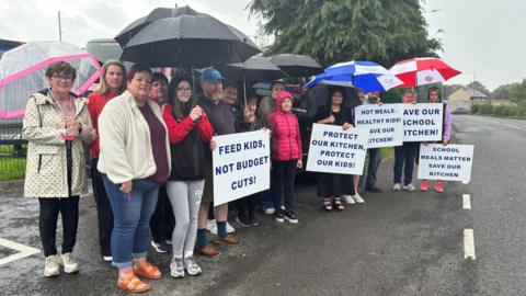 A group of adults protesting outside the school on a road. They're holding up signs reading things like 'feed kids, not budget cuts' and 'protect our kitchen'