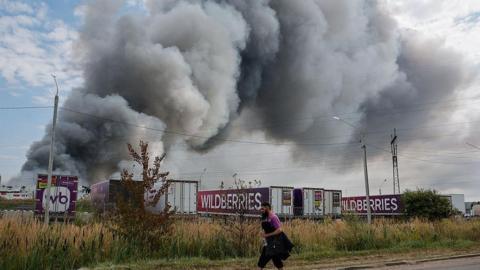 Wildberries employee walks past plumes of smoke rising above wildberries logistics complex. There are seven company lorries in front of the smoke as well as roadside shrubs