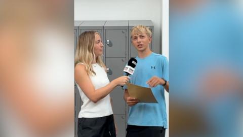 There are two people in frame standing in front of grey lockers, the person on the right hand side of the image is wearing a blue shirt and is holding a sand coloured envelope. The person on the left hand side of the image is wearing a white top and black bottoms, they are holding a microphone with bbc logos on it. Both people have blonde hair.