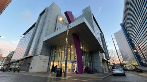 A large multi-storey modern building. There are cars parked on the pavement outside too. It is evening time and it appears it has been raining.