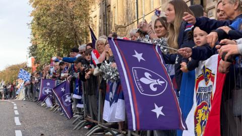 People with flags and banners line behind metal fences waiting for a bus parade. They have purple flags and are wearing red, blue and white shirts