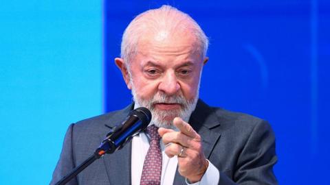 Brazilian President Luiz Inacio Lula da Silva is at a mic, seen from the elbows pointing with his left hand and he appears to be talking. He has white hair and a short white beard.