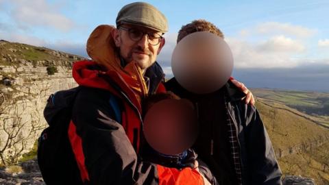 Hector Patmore wearing a red and black jacket, flat cap and glasses. He is hillwalking with two children, whose faces are blurred.