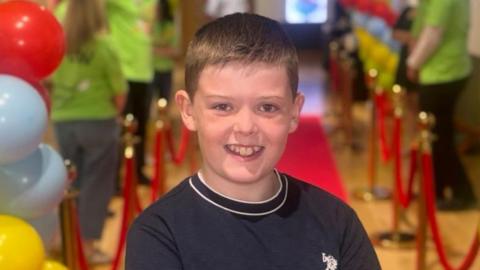 Myles is smiling at the camera. He is wearing a navy t-shirt and has brown short hair.