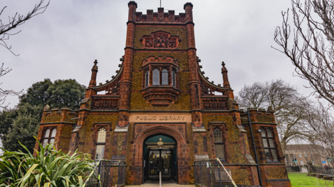 External view of old Carnegie library in King's Lynn. It is made up of a three-storey corner entrance tower with asymmetric wings, built from dark, brownish-orange carstone blocks and bright red terracotta dressings. The castle-like layout is detailed with stepped buttresses and large Gothic-style windows.