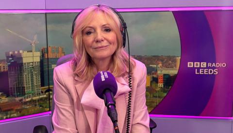 Image shows the mayor of West Yorkshire wearing a pale pink suit speaking into a BBC Radio Leeds microphone. She has headphones on and is sitting in front of an image of Leeds.