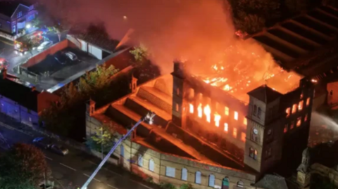 Flames and smoke from a fire engulf an old-fashioned mill buildings.