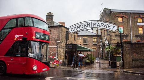 A red London bus heading to Camden Town passes by the entrance to Camden Market.