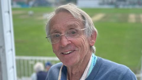 Dick Davies has grey hair and is wearing glasses. He is smiling with the cricket pitch behind him. He is standing next to the white commentary box.