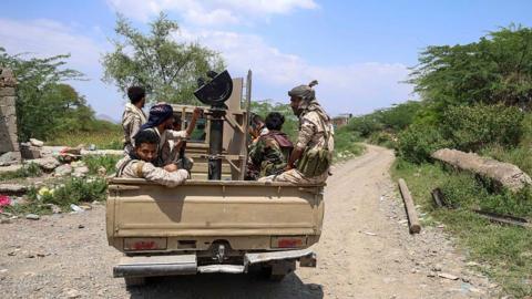 Several people ride in the back of a pick-up truck carrying mounted equipment along an unpaved road lined with trees and scrub