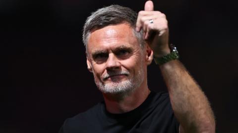 Graham Alexander, wearing a black t-shirt, smiles slightly and puts his thumbs up