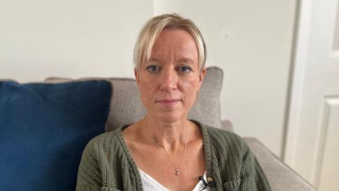 Tracy has blonde hair and is sat on her sofa looking into the camera. She has a white T-Shirt, necklace and green cardigan on.