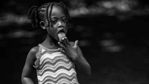 A black and white image of a young black girl with Down's syndrome licking her fingers.