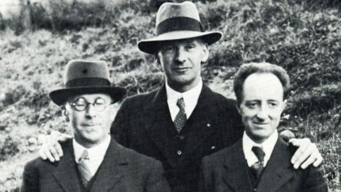 In black and white, pictured from left to right: DJ Williams, Lewis Valentine and Saunders Lewis with arms around each other. All are wearing three piece suits, Williams and Valentine are wearing fedora hats