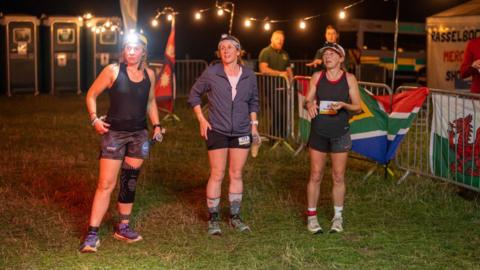 Three women standing in sports shorts and tops, and wearing caps and headbands. They are standing on grass, and there is fencing, fairy lights and portaloo toilets behind them