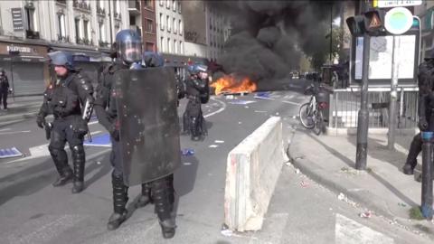 A police officer in riot gear holding a large shield stands in the street while a fire burns behind